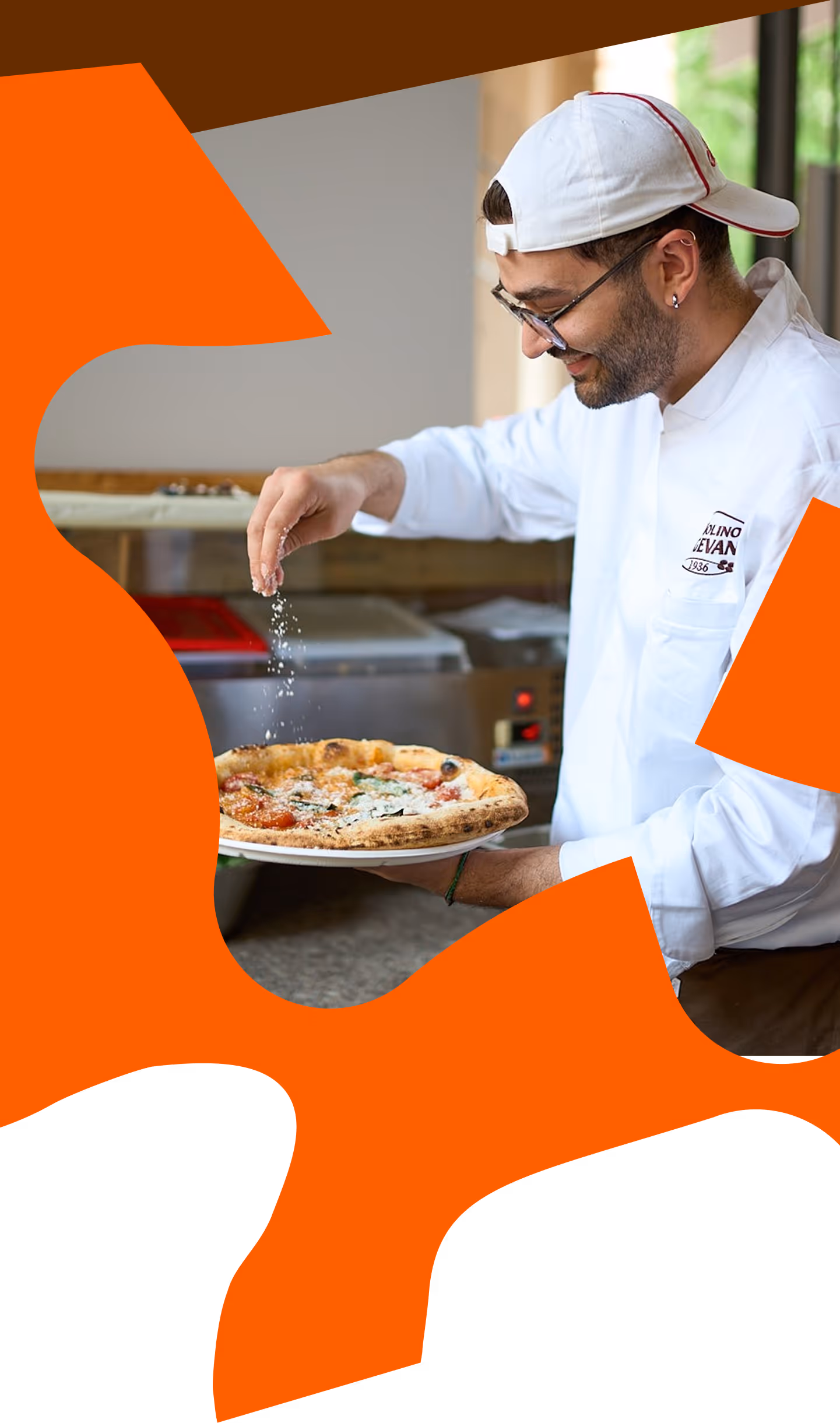 Chef wearing a white uniform and cap sprinkles seasoning on a freshly baked pizza on a plate.