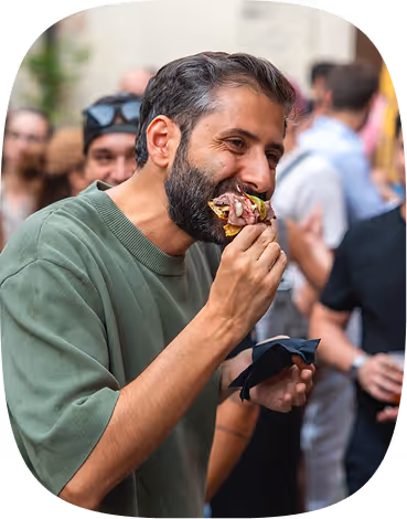Bearded man in green sweater eating a sandwich at a crowded outdoor event.
