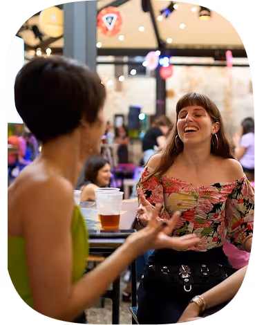 Two women laughing and talking at a table in a lively indoor cafe or bar setting.